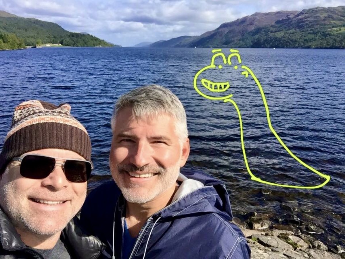 Two men smiling on the shore of Loch Ness with a cartoon Nessie drawn over the water behind them.