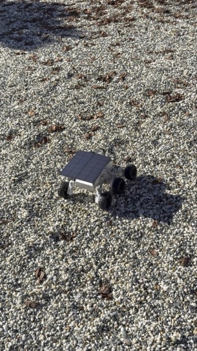 The rover from above on a wide field of pale gravel with a few fallen leaves.